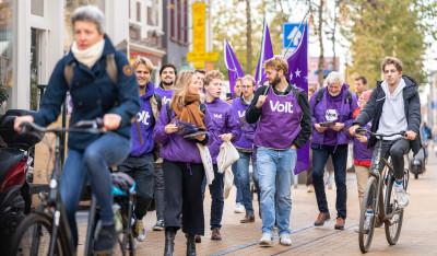 Volters lopen op straat in paarse kleding met Volt vlaggen om actie te voeren tijdens de bustour voor de 2de kamerverkiezingen 2023