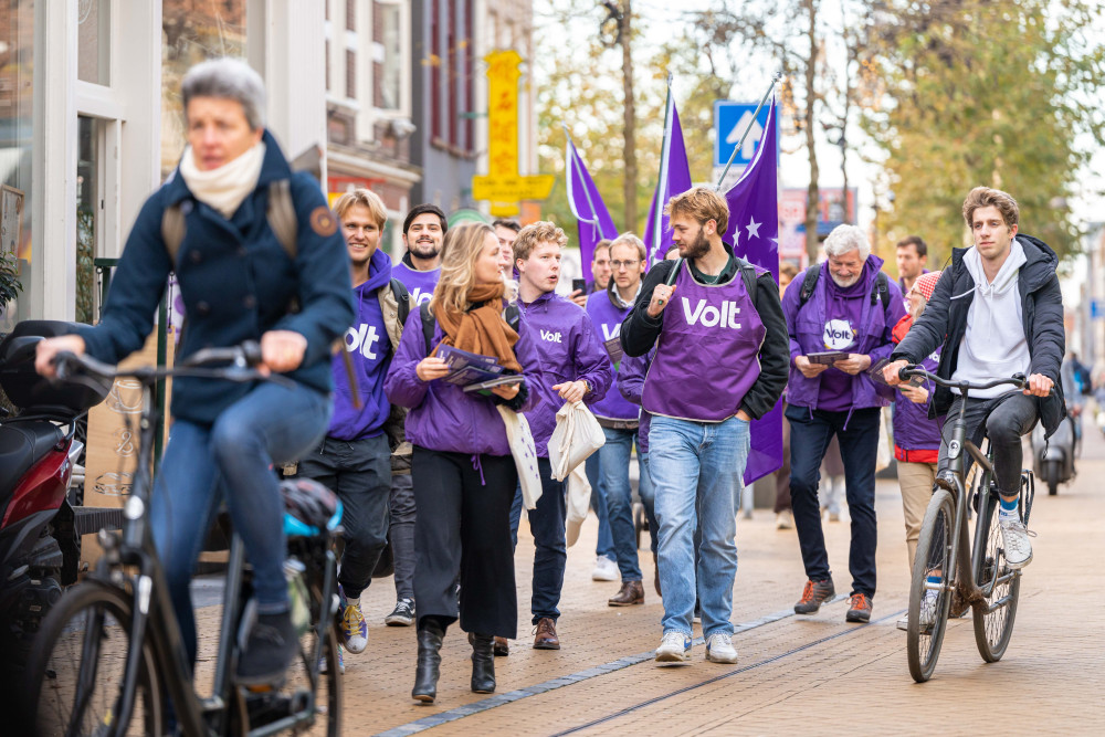 Volters lopen op straat in paarse kleding met Volt vlaggen om actie te voeren tijdens de bustour voor de 2de kamerverkiezingen 2023