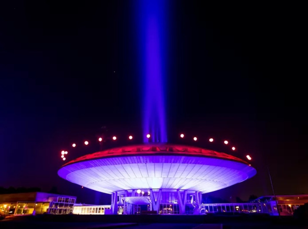 Een gebouw dat lijkt op een ufo met rood en blauwe lampen er op. Er komt een blauwe licht straal uit het gebouw.