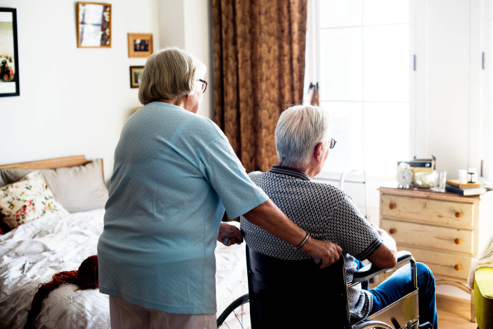 Een oudere vrouw die de rolstoel rijdt waar een oudere man in zit in een slaapkamer