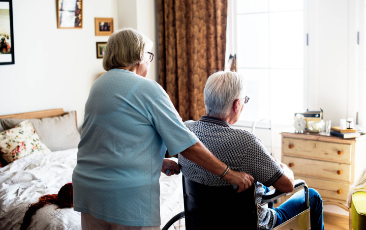 Een oudere vrouw die de rolstoel rijdt waar een oudere man in zit in een slaapkamer