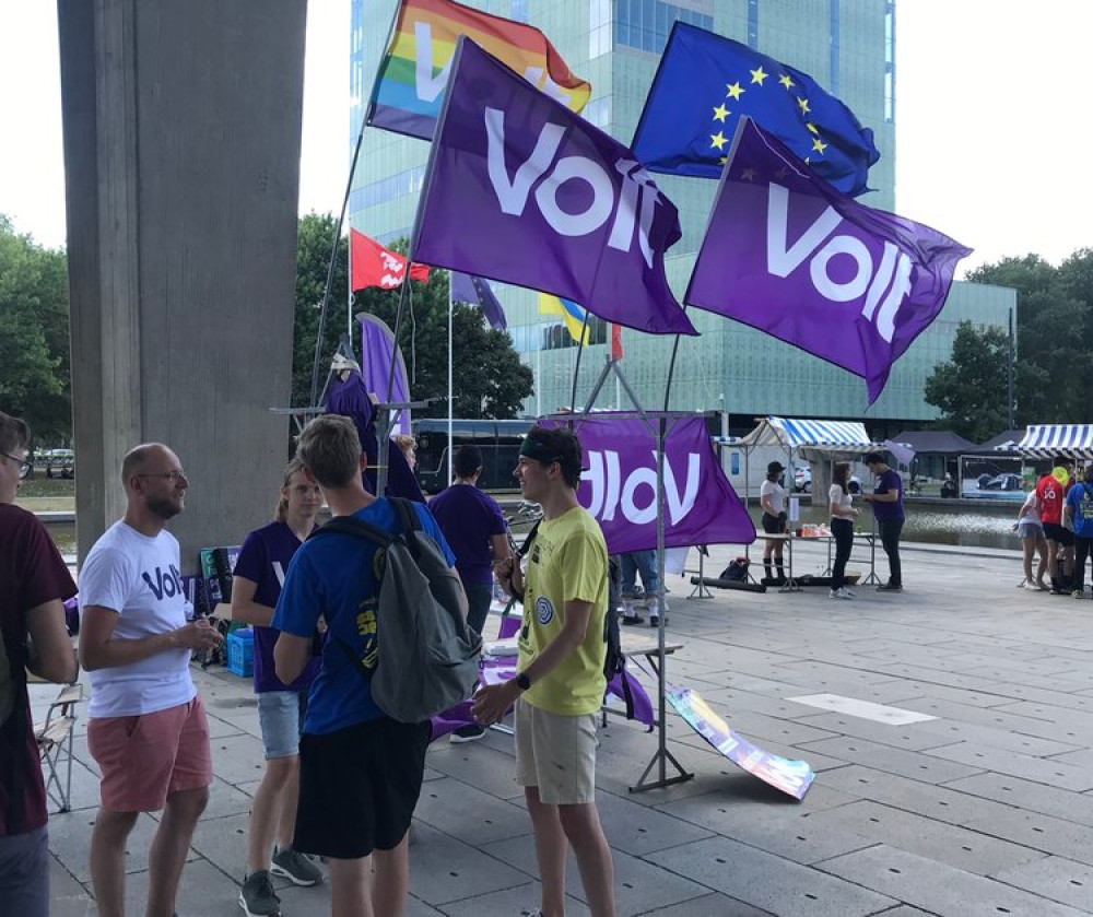 Groep mensen op een plein die aan praten zijn. Op het plein staan Volt vlaggen