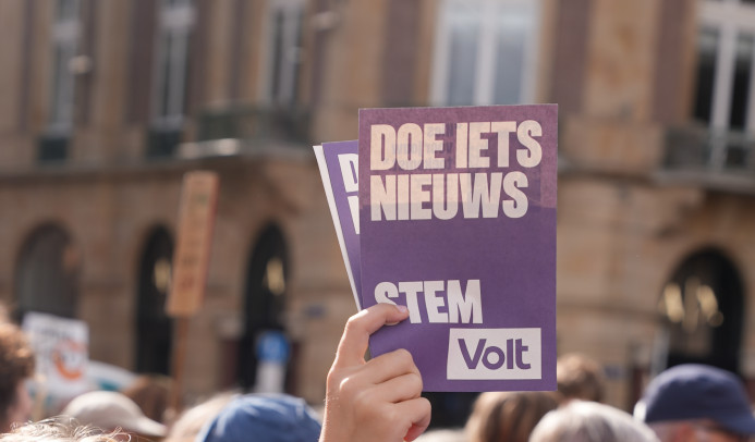 Hand houdt stapel met paarse Voltflyer omhoog voor een menigte. Op de flyer staat 'Doe iets nieuws. Stem Volt'.